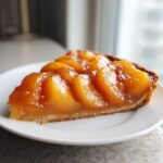 A close-up of a tart slice topped with glistening, glazed peach pie filling on a white plate.