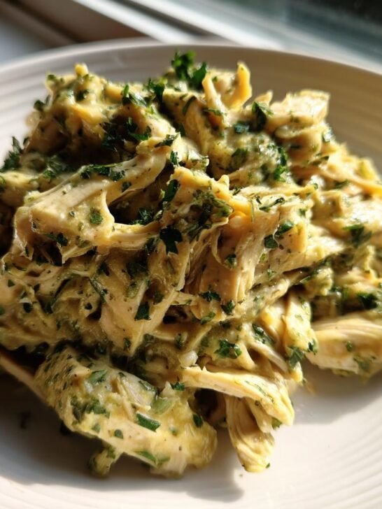 Shredded Slow Cooker Cilantro Lime Chicken coated in a creamy green sauce and fresh herbs, served on a white plate.