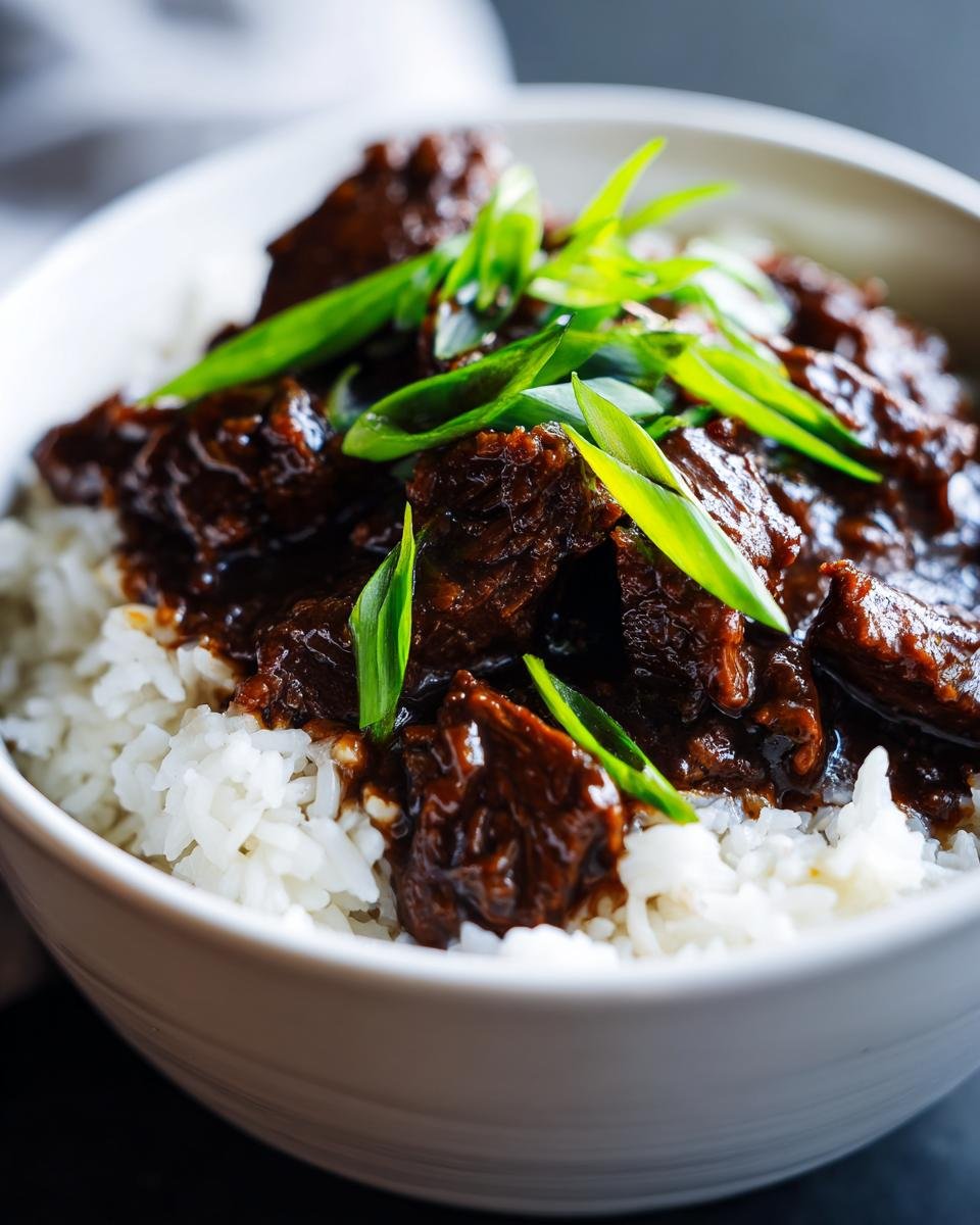 A close-up of a white bowl filled with white rice topped with rich, saucy Slow Cooker Mongolian Beef and garnished with sliced green onions.