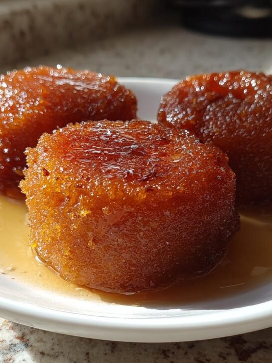 Three golden-brown, soft Gulab Jamun balls resting in a pool of sugar syrup on a white plate.