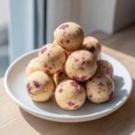 A stack of homemade Strawberry Cheesecake Protein Balls speckled with red fruit pieces, served on a white plate.