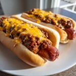 Two fully loaded Chili Cheese Dogs topped with thick chili and melted cheddar cheese on a white plate.