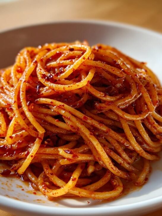 Close-up of a serving of glossy Chili Oil Pasta tossed in a vibrant red chili sauce, presented in a white bowl.