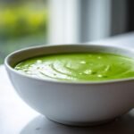 Close-up of a white bowl filled with bright, creamy Green Detox Soup sitting by a window.