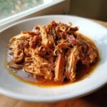 A close-up of tender, shredded Apple Cider Braised Pork Shoulder resting in its rich, savory sauce on a white plate.