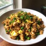 A close-up of a white bowl filled with savory Cauliflower Fried Rice With Chicken, scrambled eggs, and bright green peas, topped with scallions.