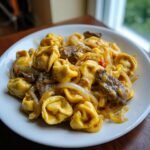 A close-up of a serving of Cheesesteak Tortellini Provolone Sauce, featuring cheese tortellini mixed with steak strips and saut&eacute;ed onions.