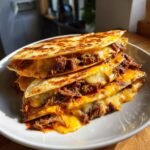 A stack of crispy, cheesy quesadillas filled with shredded meat, resembling Fave Birria Tacos, oozing melted cheese.