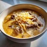 A close-up of a bowl of rich, creamy Philly Cheese Steak Soup topped generously with shredded and melting cheese.