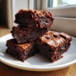 A stack of four rich, dark Fudgy Chewy Browkies piled on a white plate, showing their dense, moist texture.