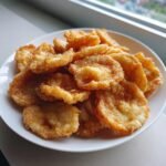 A pile of crispy, golden brown Fried Mushrooms served on a white plate near a window.