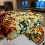 A hand holding a thick slice of Greek Roasted Garlic Spinach White Pizza with melted, browned cheese.