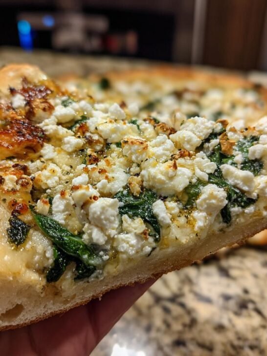 A hand holding up a thick slice of Greek Roasted Garlic Spinach White Pizza topped with feta and spinach.