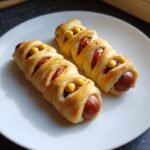 Two golden-brown Halloween Air Fryer Mummy Hot Dogs wrapped in dough strips, decorated with mustard eyes, served on a white plate.