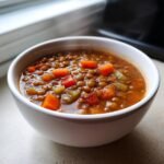 A close-up of a white bowl filled with rich Lentil Vegetable Soup featuring visible lentils, carrots, and celery.