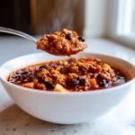 A spoonful of steaming Hearty Turkey Apple Chili being lifted from a white bowl, showing ground turkey, beans, and apple chunks.