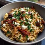 Close-up of a bowl filled with High Protein Mediterranean Chicken Orzo, topped with feta, sun-dried tomatoes, and parsley.