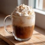 A clear glass mug filled with a Homemade Pumpkin Spice Latte, topped high with whipped cream and a sprinkle of spice.