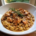 A close-up of creamy Marsala Chicken Orzo Pasta topped with a fresh rosemary sprig in a white bowl.