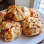 A close-up of several golden-brown Monster Chicken Bacon Ranch Bombs piled on a white plate, topped with melted cheese and bacon bits.
