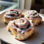 Close-up of three fluffy Pumpkin Cinnamon Rolls Browned Butter drizzled with thick white icing on a white plate.