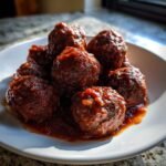 A close-up of several juicy Red Wine Braised Meatballs coated in a rich, dark sauce, served on a white plate.
