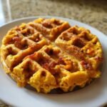 A close-up of a golden, circular Savory Cornbread Waffles, studded with bits of bacon or cheese, served on a white plate.