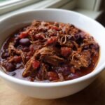 A close-up of a hearty bowl of Short Rib Chorizo Chili, featuring shredded meat and kidney beans in a rich, dark red sauce.
