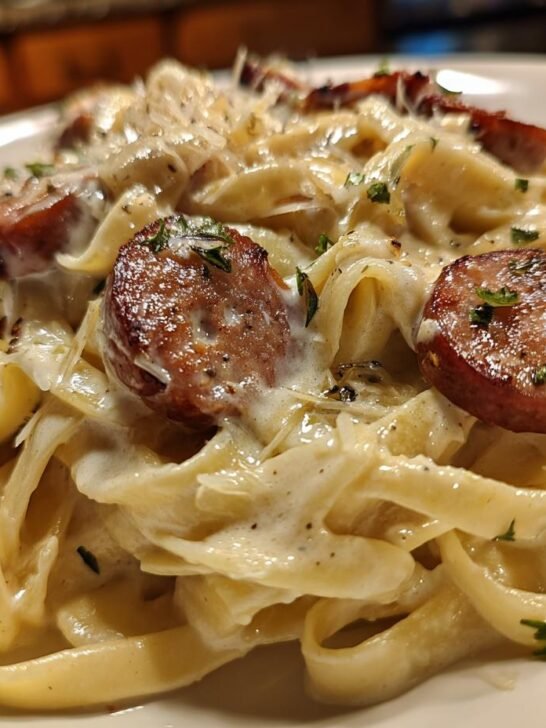 Close-up of creamy fettuccine pasta tossed in Alfredo sauce with slices of browned smoked sausage, garnished with parsley.
