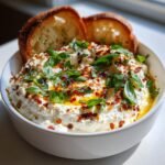 A bowl of creamy Spicy Ricotta Dip topped with fresh basil, chili flakes, and olive oil, served with two slices of toasted bread.