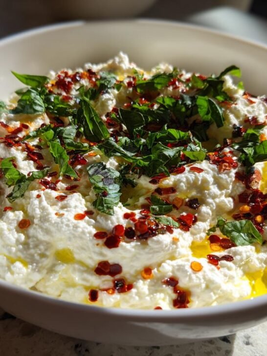 Close-up of a white bowl filled with creamy Spicy Ricotta Dip, drizzled with olive oil and topped with red pepper flakes and fresh basil.
