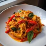 A close-up of shredded Spicy Thai Red Curry Pot Roast Chicken mixed with red bell peppers and garnished with fresh basil leaves.