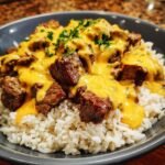 A close-up shot of Steak Queso Rice featuring chunks of steak smothered in bright yellow queso sauce over white rice, garnished with parsley.