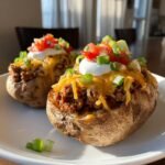 Two fully loaded Taco Stuffed Baked Potatoes topped with seasoned beef, melted cheese, sour cream, tomatoes, and green onions.