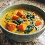 Close-up of a bowl filled with vibrant orange Vegetable Coconut Stew featuring chunks of sweet potato and wilted spinach.
